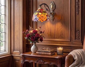 May include: A wall-mounted lamp with a stained glass shade featuring orange, red, and purple floral patterns. The lamp has an ornate bronze-toned metal arm and base. It is positioned above a wooden side table holding a vase of roses.
