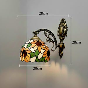 May include: A wall-mounted Tiffany-style lamp with a sunflower design. The lamp shade is 20 cm wide and 28 cm high, featuring yellow, orange, green, and white glass. The fixture has a bronze finish.