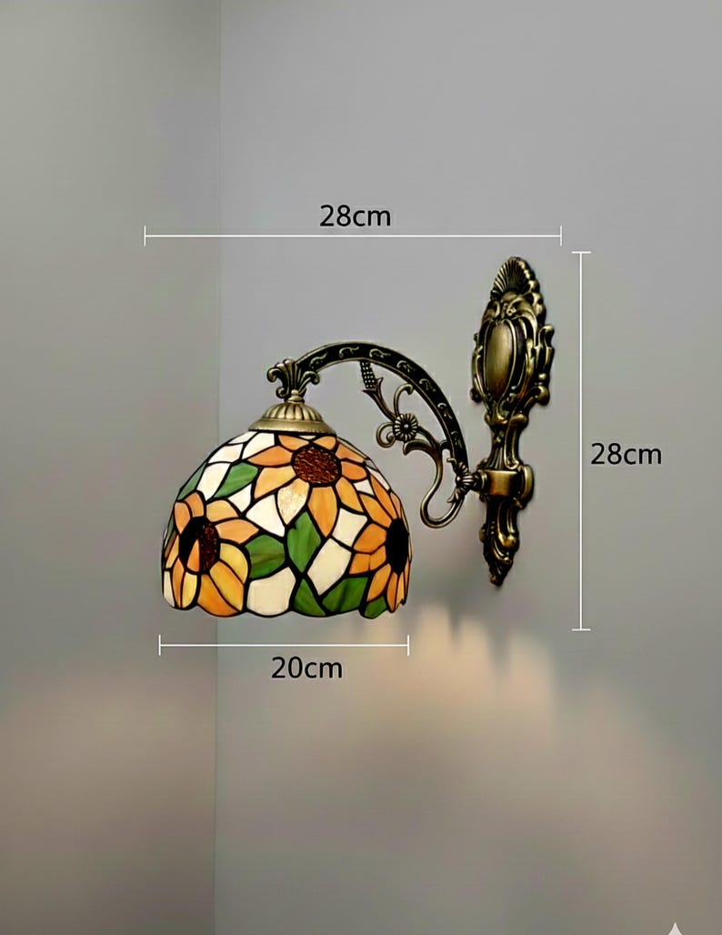 May include: A wall-mounted Tiffany-style lamp with a sunflower design. The lamp shade is 20 cm wide and 28 cm high, featuring yellow, orange, green, and white glass. The fixture has a bronze finish.