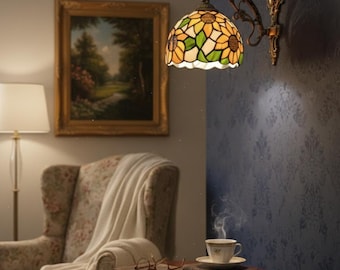 May include: A wall-mounted Tiffany-style lamp with a sunflower design. The lamp features a stained glass shade with yellow, green, and white panels. The lamp is mounted on a dark metal arm and bracket. A painting, armchair, and side table with a cup of tea are also visible.