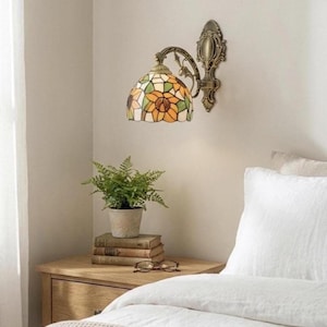 Könnte beinhalten: Eine Wandleuchte mit einem Lampenschirm aus Buntglas mit einem Sonnenblumenmuster in Gelb, Orange und Grün. Die Lampe hat einen bronzefarbenen Metallrahmen und ist an einer Wand befestigt. Eine kleine Topfpflanze und Bücher stehen auf einem Nachttisch aus Holz.
