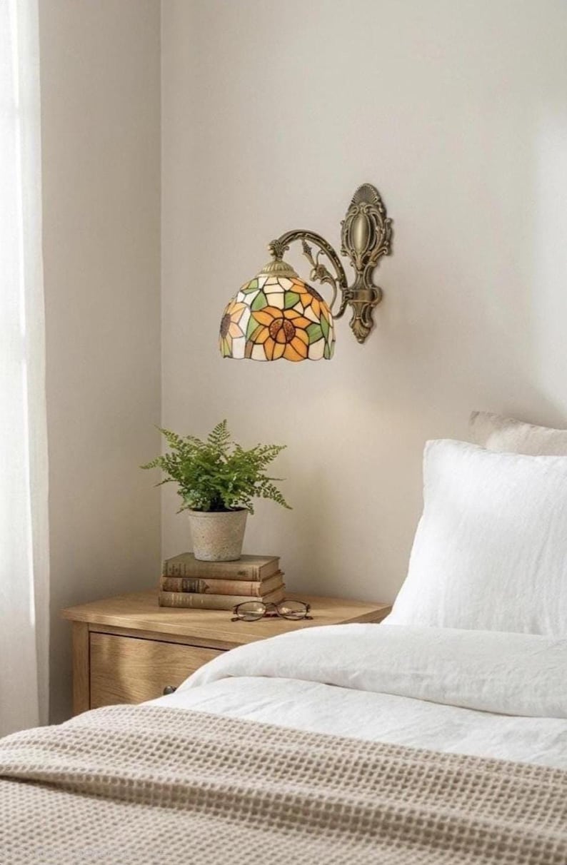 May include: A wall-mounted lamp with a stained glass shade featuring a sunflower design in yellow, orange, and green. The lamp has a bronze-colored metal frame and is mounted on a light-colored wall. A small potted plant and books sit on a wooden nightstand.