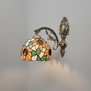 May include: A wall-mounted lamp with a stained-glass shade featuring a sunflower design in shades of yellow, orange, green, and brown. The lamp has a bronze-colored metal frame and decorative accents.