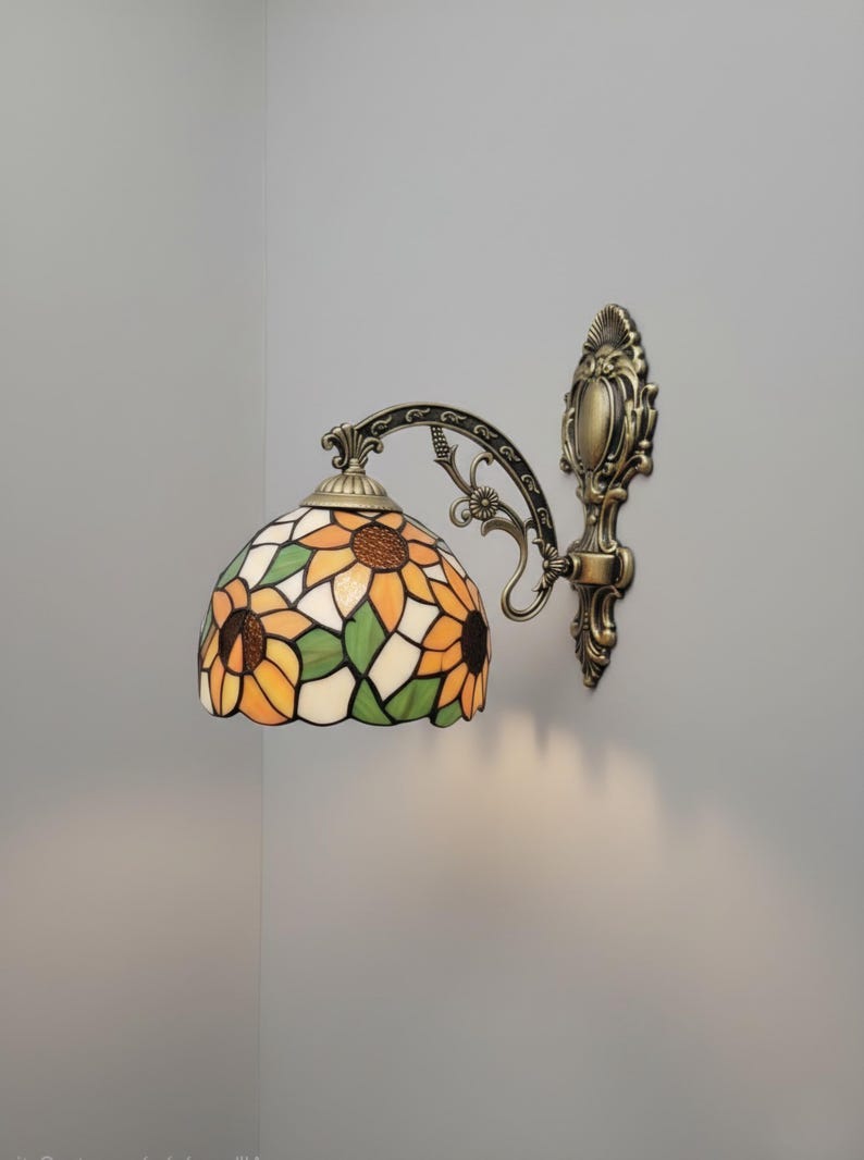 May include: A wall-mounted lamp with a stained-glass shade featuring a sunflower design in shades of yellow, orange, green, and brown. The lamp has a bronze-colored metal frame and decorative accents.