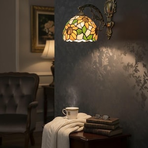 May include: A wall-mounted lamp with a stained glass shade featuring a sunflower design in yellow, green, and white. The lamp has a bronze-colored metal frame. A small table holds a cup of tea, books, and glasses.