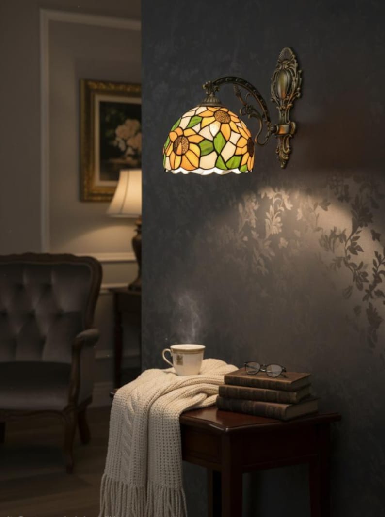 May include: A wall-mounted lamp with a stained glass shade featuring a sunflower design in yellow, green, and white. The lamp has a bronze-colored metal frame. A small table holds a cup of tea, books, and glasses.