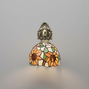 May include: A wall-mounted lamp with a stained-glass shade featuring a sunflower design. The shade has orange, yellow, green, and white glass pieces. The lamp's base and mounting hardware are bronze-colored.
