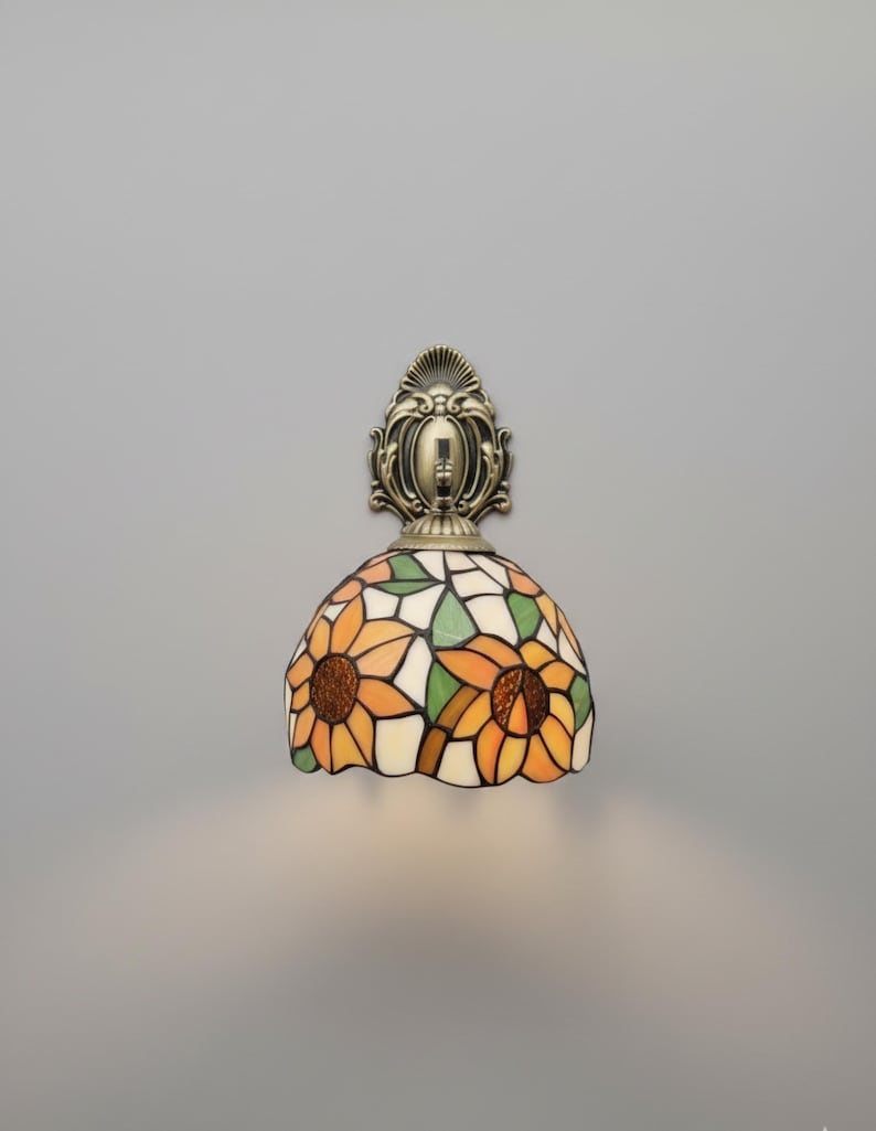 May include: A wall-mounted lamp with a stained-glass shade featuring a sunflower design. The shade has orange, yellow, green, and white glass pieces. The lamp's base and mounting hardware are bronze-colored.