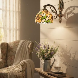 May include: A wall-mounted lamp with a stained glass shade featuring a sunflower design in yellow, green, and white. The lamp has a bronze-colored metal frame. A small wooden shelf holds a watering can, flowers, a teacup, and a book.