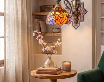 May include: A wall-mounted lamp featuring a stained glass shade with a floral rose pattern in shades of orange, red, yellow, and purple. The lamp has an ornate, dark metal fixture attached to the wall, providing illumination to the surrounding area.