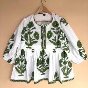 May include: A white long-sleeve blouse featuring a green floral block print pattern. The garment has a round neckline with a central lace-up closure, a gathered waistline, and elasticized cuffs. The fabric is white with green botanical motifs.