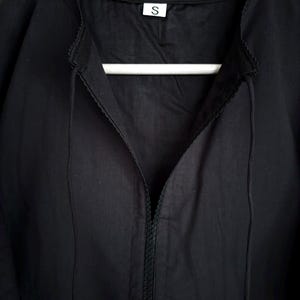 May include: A black dress with a V-neckline and a small, decorative trim along the collar and neckline. The dress is hanging on a white hanger. A small white tag with the letter "S" is visible.