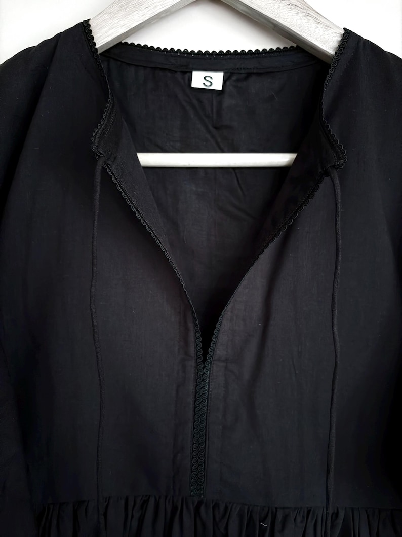 May include: A black dress with a V-neckline and a small, decorative trim along the collar and neckline. The dress is hanging on a white hanger. A small white tag with the letter "S" is visible.