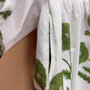Puede incluir: Primer plano de tela blanca adornada con un estampado floral verde. El dise&ntilde;o presenta grandes flores y hojas verdes estilizadas. La tela parece ser de algod&oacute;n o lino ligero, con un detalle de bolsillo visible.