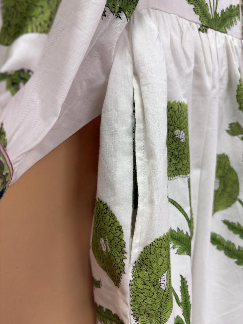 Puede incluir: Primer plano de tela blanca adornada con un estampado floral verde. El dise&ntilde;o presenta grandes flores y hojas verdes estilizadas. La tela parece ser de algod&oacute;n o lino ligero, con un detalle de bolsillo visible.