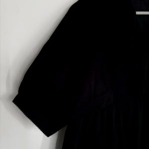 May include: Black short-sleeved dress with a V-neckline. The dress features a gathered waist and scalloped edges on the sleeves. The dress is hanging on a white background.