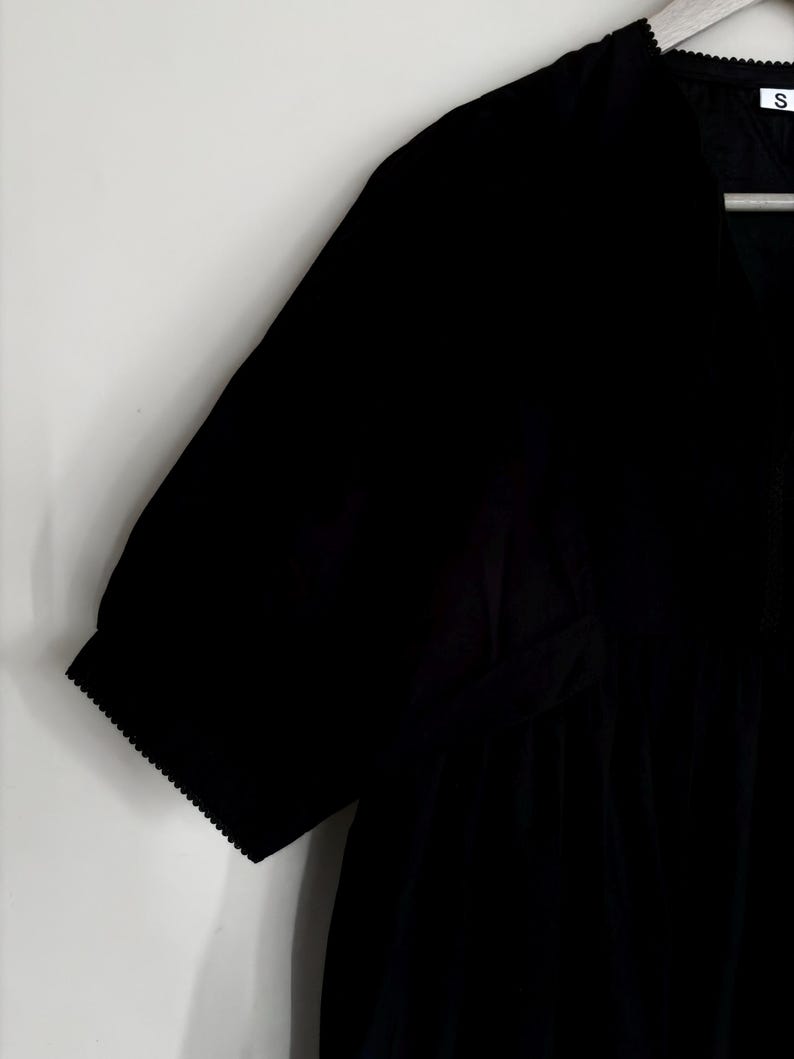 May include: Black short-sleeved dress with a V-neckline. The dress features a gathered waist and scalloped edges on the sleeves. The dress is hanging on a white background.