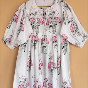 May include: White tunic dress with a floral print in pink and green. The dress features a round neckline, short sleeves, and a tie detail at the waist. The floral pattern is repeated across the fabric, creating a cohesive design.