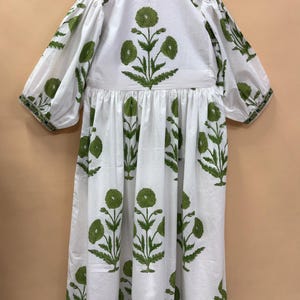 Puede incluir: Vestido blanco con estampado floral verde. El vestido presenta un cuello redondo, mangas cortas y cintura fruncida. El dise&ntilde;o floral verde se repite, con ribete verde en las mangas y el dobladillo.