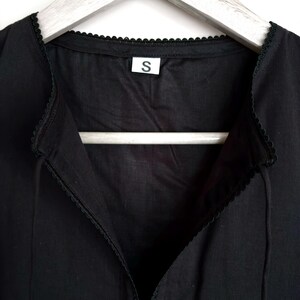 May include: Close-up of a black garment with a V-neck design and delicate lace trim along the neckline and edges. A small white tag with the letter "S" is visible. The garment is hanging on a wooden hanger.