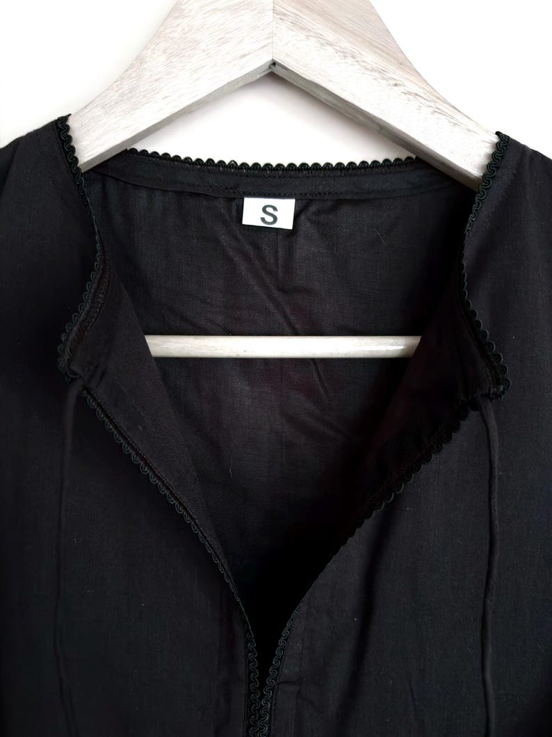 May include: Close-up of a black garment with a V-neck design and delicate lace trim along the neckline and edges. A small white tag with the letter "S" is visible. The garment is hanging on a wooden hanger.