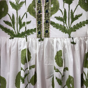 Puede incluir: Vestido blanco con estampado floral verde. El vestido presenta un escote en V con detalles bordados en verde y azul. La tela est&aacute; fruncida en la cintura y el vestido tiene una silueta suelta y fluida. El dise&ntilde;o floral incluye flores y hojas grandes y estilizadas.