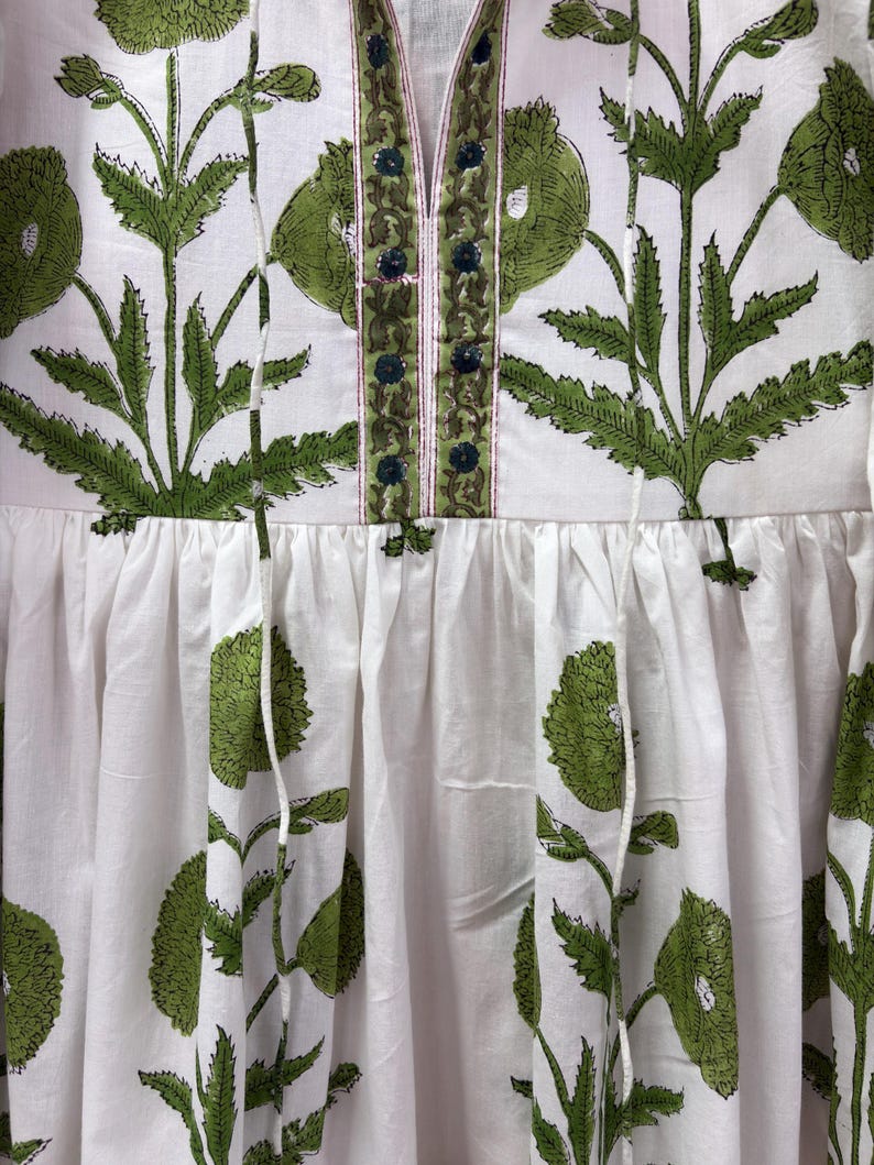 Puede incluir: Vestido blanco con estampado floral verde. El vestido presenta un escote en V con detalles bordados en verde y azul. La tela est&aacute; fruncida en la cintura y el vestido tiene una silueta suelta y fluida. El dise&ntilde;o floral incluye flores y hojas grandes y estilizadas.
