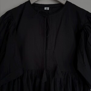 May include: Black dress with a round neckline, short puff sleeves, and a gathered skirt. The dress has a tie detail at the neckline and is hanging on a wooden hanger. The tag on the dress has the letter "S".