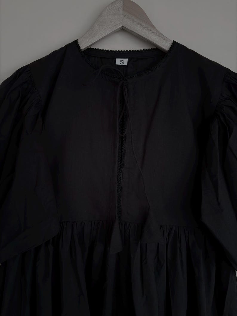 May include: Black dress with a round neckline, short puff sleeves, and a gathered skirt. The dress has a tie detail at the neckline and is hanging on a wooden hanger. The tag on the dress has the letter "S".