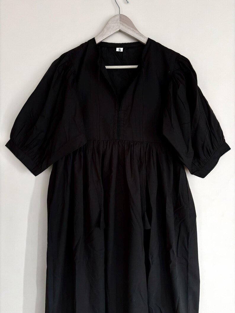 May include: A black dress with short, puffed sleeves and a gathered waist. The dress has a V-neckline and a small white tag at the neck. The dress is hanging on a wooden hanger.
