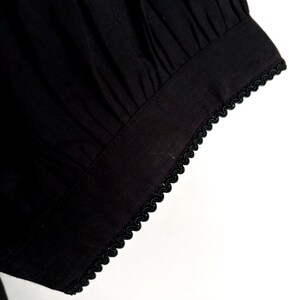 May include: Close-up of a black fabric sleeve with a gathered design. The cuff is trimmed with a delicate, scalloped black trim. The fabric appears to be a lightweight material, possibly cotton or linen.