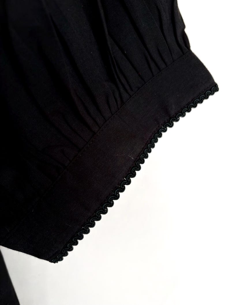 May include: Close-up of a black fabric sleeve with a gathered design. The cuff is trimmed with a delicate, scalloped black trim. The fabric appears to be a lightweight material, possibly cotton or linen.
