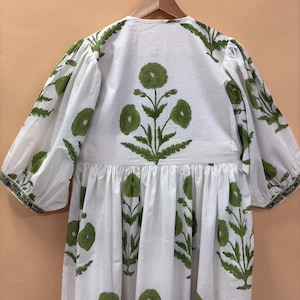 Puede incluir: Vestido blanco con estampado floral verde. El vestido presenta un cuello en V, mangas tres cuartos y cintura fruncida. Las mangas son abullonadas. El vestido est&aacute; colgado en una percha de madera.