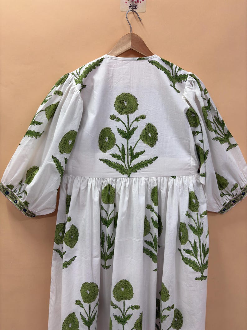 Puede incluir: Vestido blanco con estampado floral verde. El vestido presenta un cuello en V, mangas tres cuartos y cintura fruncida. Las mangas son abullonadas. El vestido est&aacute; colgado en una percha de madera.