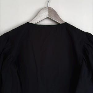 May include: Black blouse with a scalloped neckline and long, gathered sleeves. The garment is hanging on a wooden hanger against a white background. The blouse has a ruffled hemline.