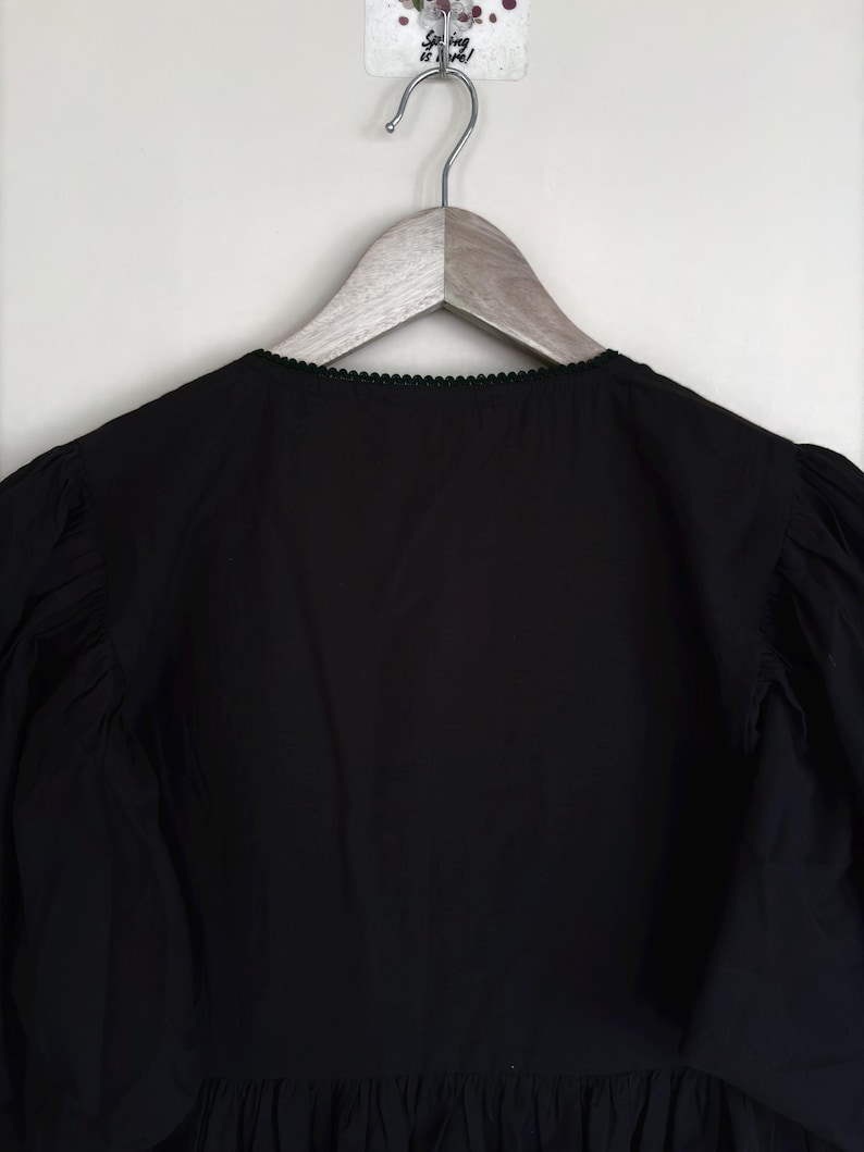 May include: Black blouse with a scalloped neckline and long, gathered sleeves. The garment is hanging on a wooden hanger against a white background. The blouse has a ruffled hemline.