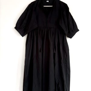 May include: A black midi dress with a V-neckline and short, puffed sleeves. The dress features a gathered waist and a flowing skirt. It is hanging on a wooden hanger against a white background. The hemline and sleeve cuffs have a delicate trim.