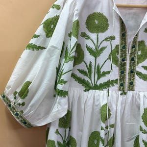 Puede incluir: Vestido blanco con estampado floral verde. El vestido presenta mangas cortas, cuello en V y cintura fruncida. El estampado floral verde se repite por todo el vestido, con un borde detallado en las mangas y el escote. El vestido est&aacute; colgado en una percha de madera.