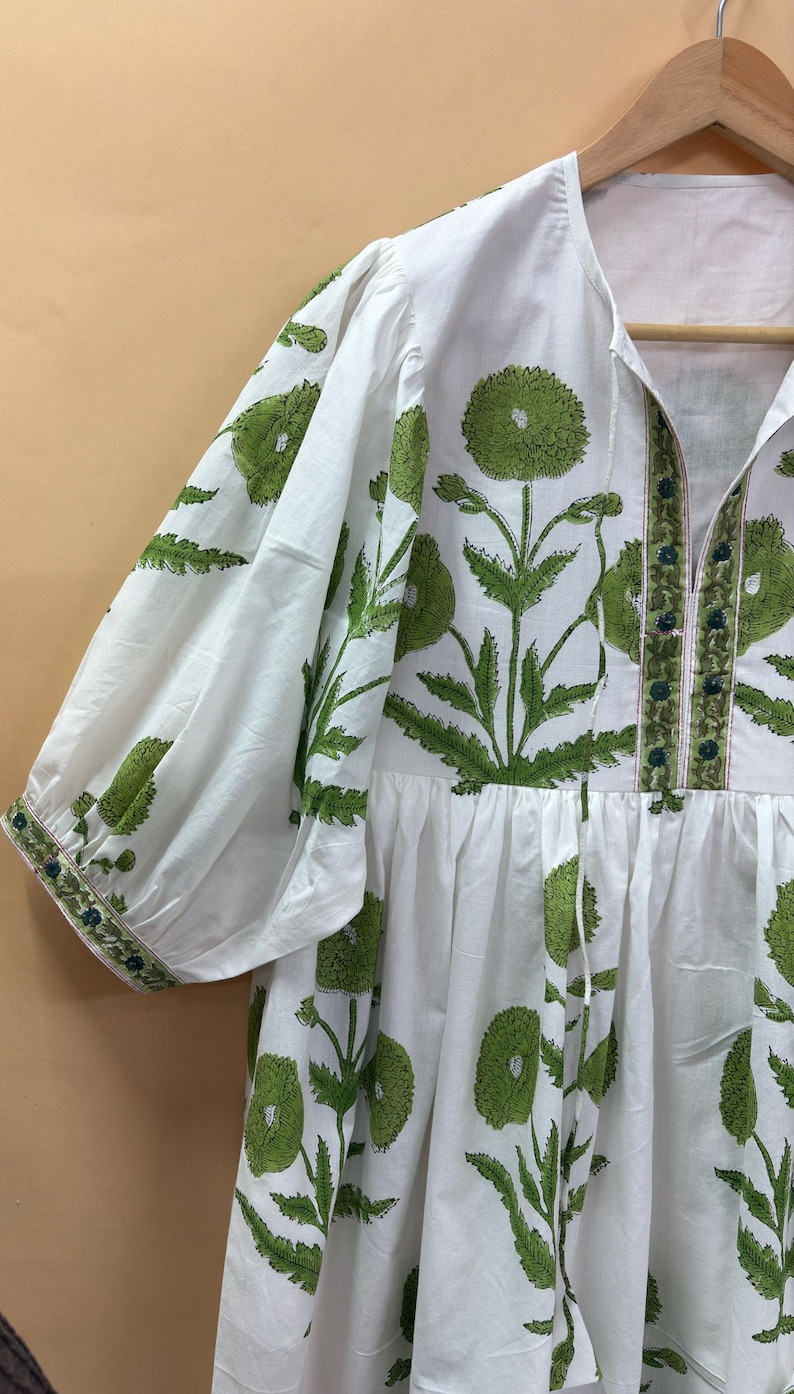 Puede incluir: Vestido blanco con estampado floral verde. El vestido presenta mangas cortas, cuello en V y cintura fruncida. El estampado floral verde se repite por todo el vestido, con un borde detallado en las mangas y el escote. El vestido est&aacute; colgado en una percha de madera.