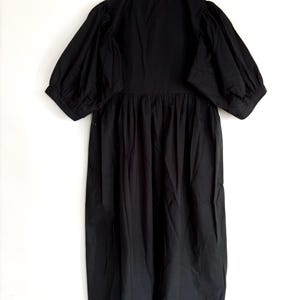May include: A black dress with a round neckline and short, puffed sleeves. The dress has a gathered waist and a long, flowing skirt. The hemline and neckline are trimmed with a delicate, scalloped edge. The dress is hanging on a wooden hanger.