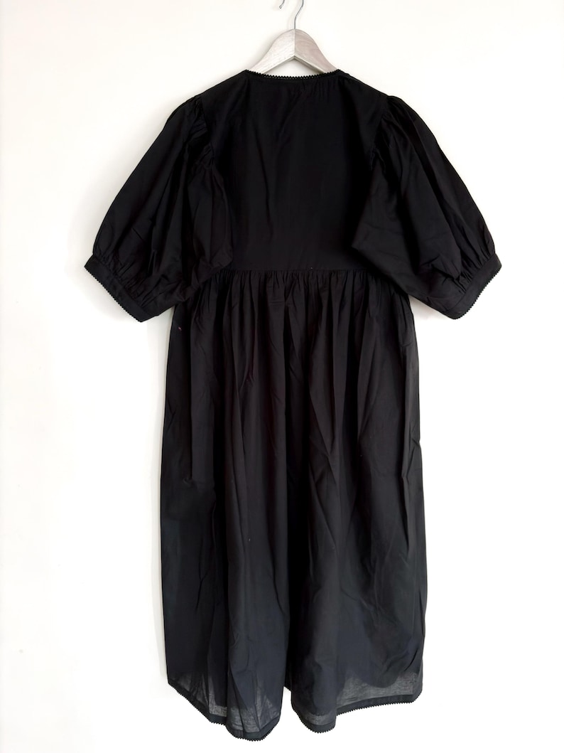 May include: A black dress with a round neckline and short, puffed sleeves. The dress has a gathered waist and a long, flowing skirt. The hemline and neckline are trimmed with a delicate, scalloped edge. The dress is hanging on a wooden hanger.