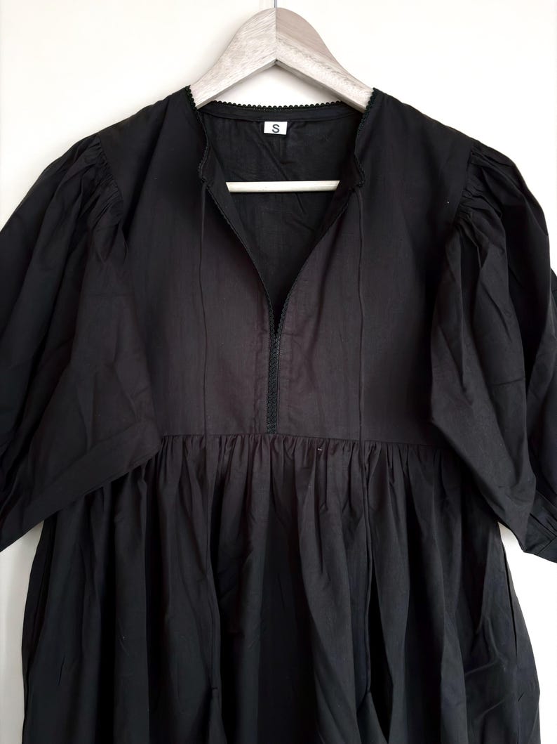 May include: A black dress with a V-neckline and short, puffed sleeves. The dress features a gathered waist and a small, decorative trim around the neckline. The dress is hanging on a wooden hanger.