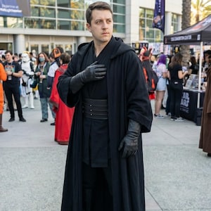 以下が含まれることがあります： 黒いフード付きローブ、手袋、ベルトを身につけた人が屋外でポーズをとっています。 その人は、背景にコスチュームを着た他の人々やテントがあるコンベンションにいます。 その人はコスチュームを着ています。