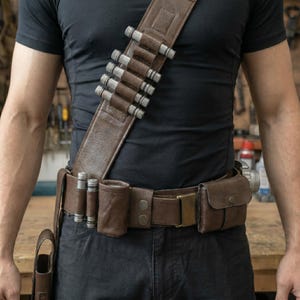 May include: Brown leather bandolier and belt set with silver-colored cylindrical accents. The bandolier crosses the chest, and the belt features pouches and a brass buckle. The set is designed for carrying items.