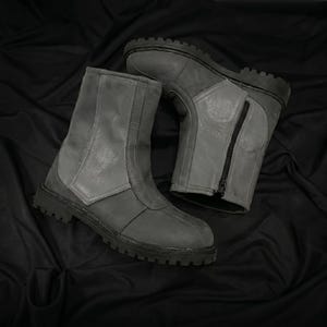 May include: A pair of gray leather boots with a rugged, treaded sole. The boots have a mid-calf height, a front zipper, and a panel design. The boots are displayed on a black fabric background.