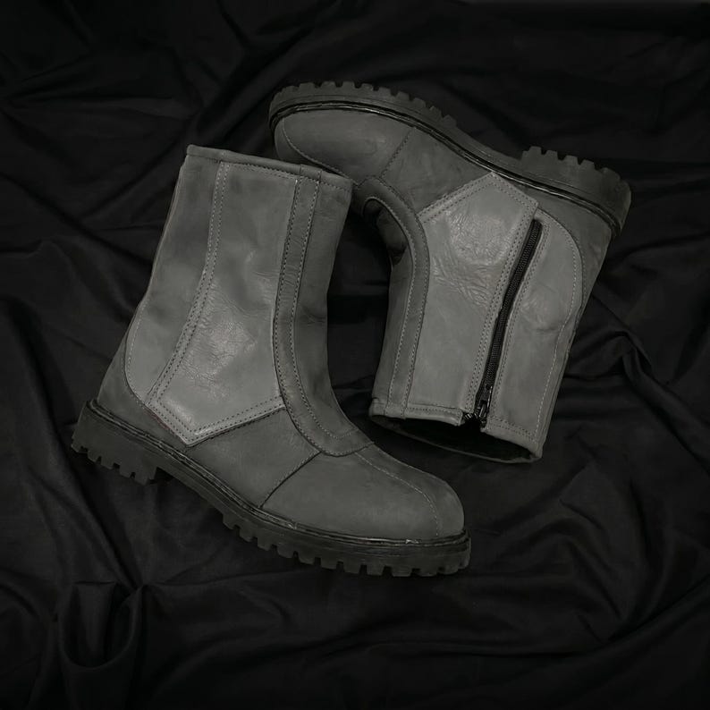 May include: A pair of gray leather boots with a rugged, treaded sole. The boots have a mid-calf height, a front zipper, and a panel design. The boots are displayed on a black fabric background.