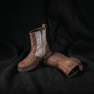 K&ouml;nnte beinhalten: Ein Paar braune Lederstiefel mit robustem Design. Die Stiefel haben einen Rei&szlig;verschluss und ein graues Panel an der Seite. Die Sohlen sind dick und strukturiert. Die Stiefel werden auf einer dunklen, strukturierten Oberfl&auml;che pr&auml;sentiert.