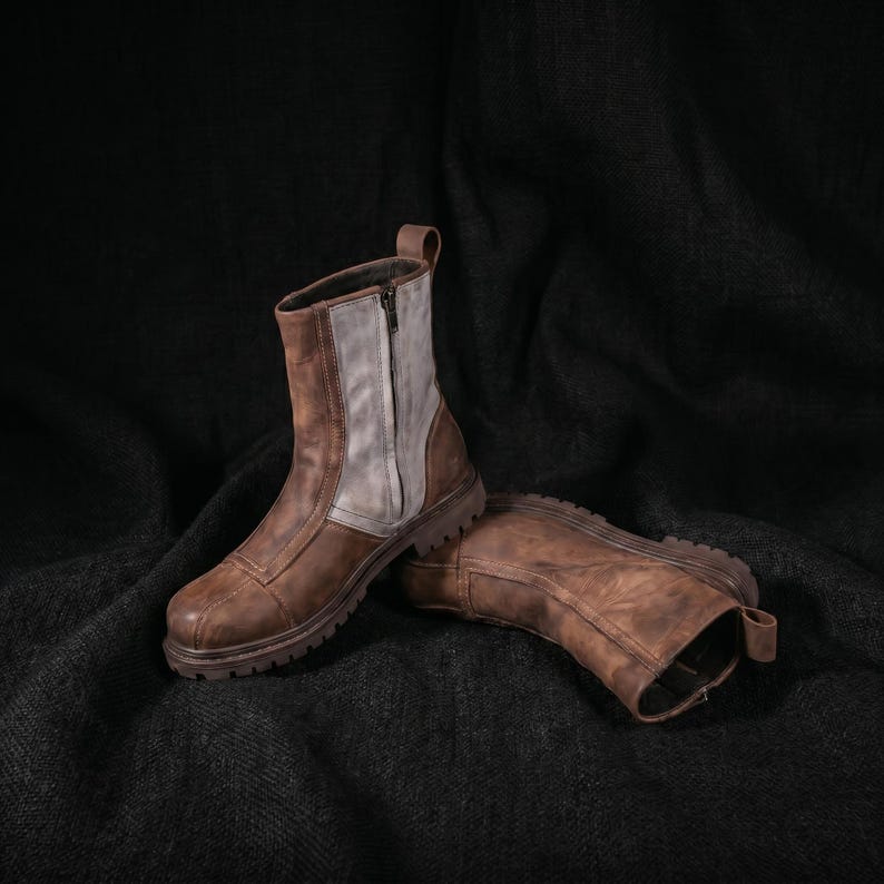 K&ouml;nnte beinhalten: Ein Paar braune Lederstiefel mit robustem Design. Die Stiefel haben einen Rei&szlig;verschluss und ein graues Panel an der Seite. Die Sohlen sind dick und strukturiert. Die Stiefel werden auf einer dunklen, strukturierten Oberfl&auml;che pr&auml;sentiert.