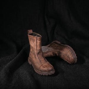K&ouml;nnte beinhalten: Zwei braune Lederstiefel mit einem Used-Look. Die Stiefel haben eine mittlere Wadenh&ouml;he, einen Rei&szlig;verschluss und eine dicke, strukturierte Sohle. Das Leder weist sichtbare Maserung und ein abgenutztes Aussehen auf, was auf Haltbarkeit und Vintage-&Auml;sthetik hindeutet.