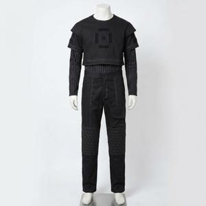 Op de afbeelding: Zwart ensemble op een mannequin. Het bevat een crop top met korte mouwen en een geometrisch ontwerp, een gestreept shirt met lange mouwen en een zwarte broek met gewatteerde kniepanelen en zakdetails.